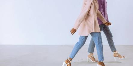 Two women in pastel shirts, blue jeans, and white heeled sandals walking side by side indoors.