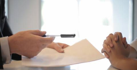 Two people sitting across from each other a desk, only their hands are visable. The one on the left is holding a contract and a pen.