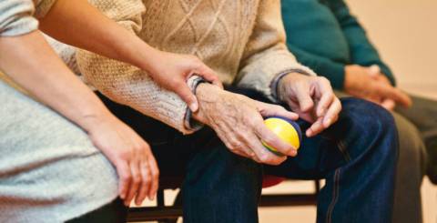 Three elderly people sitting on a bench. The man in the middle is holding a ball while the woman on the far right holds his forearm.