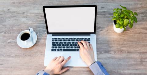 Macbook open on a desk with a black coffee to the left and a small green plant to the right. A man has both hands out and is typing on on the laptop.