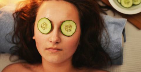 Woman laying down with cucumber slices over her eyes and a bowl of sliced cucumbers by her head on the right hand side.