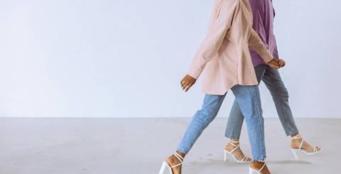 Stylish Women Walking in Casual Chic Outfits Two women in pastel shirts, blue jeans, and white heeled sandals walking side by side indoors.